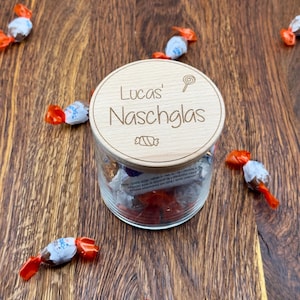 May include: A wooden jar with a lid that says "Lucas' Naschglas" with a candy cane and a swirl design. The jar is filled with wrapped candies.