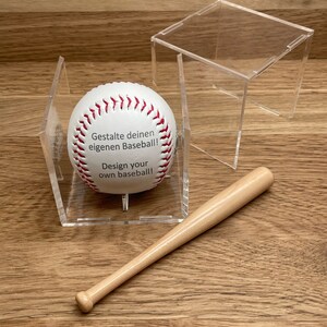 Puede incluir: Una pelota de béisbol blanca con costuras rojas y el texto "Gestalte deinen eigenen Baseball! Design your own baseball!" en una vitrina de acrílico transparente con un bate de béisbol de madera.