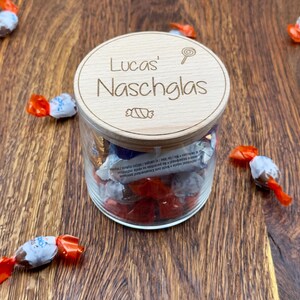 May include: A wooden jar with a lid that says "Lucas' Naschglas" with a candy cane and a lollipop graphic. The jar is filled with wrapped chocolates.