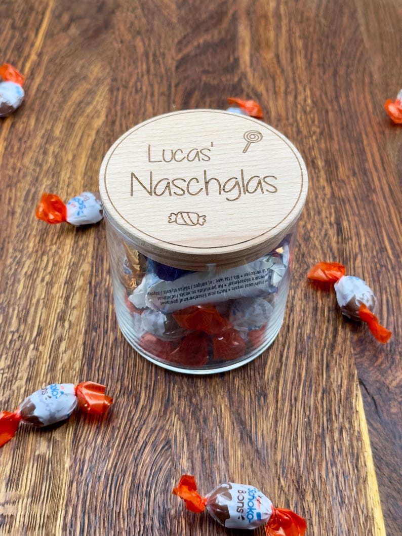 May include: A wooden jar with a lid that says "Lucas' Naschglas" with a candy cane and a lollipop graphic. The jar is filled with wrapped chocolates.