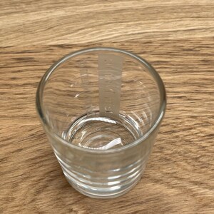 Op de afbeelding: Een helder glazen shotglas met een verhoogd, getextureerd ontwerp rond de bodem. Het glas heeft een wit label met de tekst "AUSTRIA" erop gedrukt.