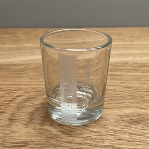 Op de afbeelding: Een helder glazen shotglas met het woord "Dennys" erop gegraveerd.