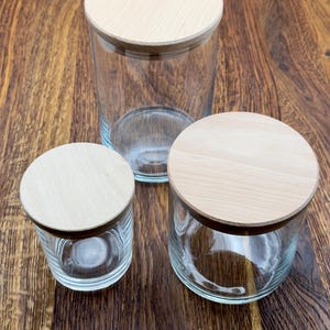 May include: Three glass jars with wooden lids. The jars are clear glass and the lids are a light wood color. The jars are different sizes, with the largest jar in the back and the smallest jar in the front.
