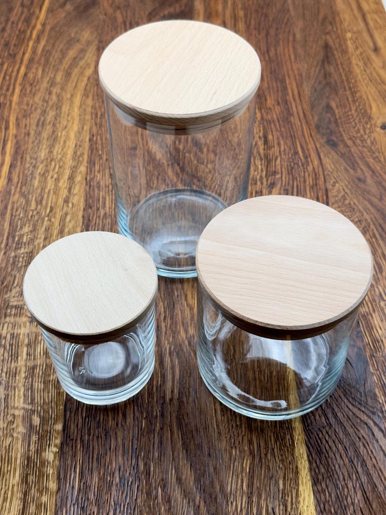 May include: Three glass jars with wooden lids. The jars are clear glass and the lids are a light wood color. The jars are different sizes, with the largest jar in the back and the smallest jar in the front.