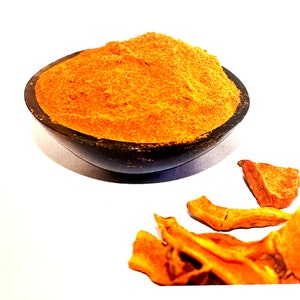 Puede incluir: Un pequeño cuenco de madera lleno de polvo de cúrcuma de color naranja brillante. Varias piezas de raíz de cúrcuma seca están esparcidas alrededor del cuenco.