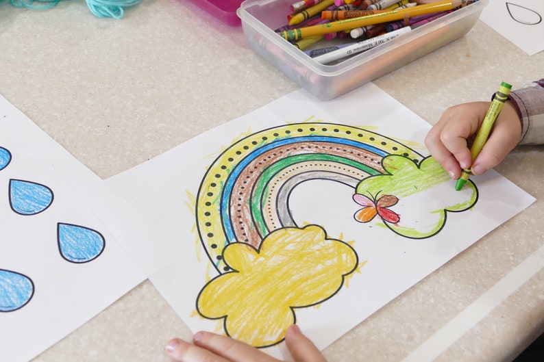 May include: A child is colouring a rainbow with a green crayon. The rainbow is drawn on a white sheet of paper with a yellow cloud and a butterfly. The rainbow is coloured in brown, green, blue, and yellow.