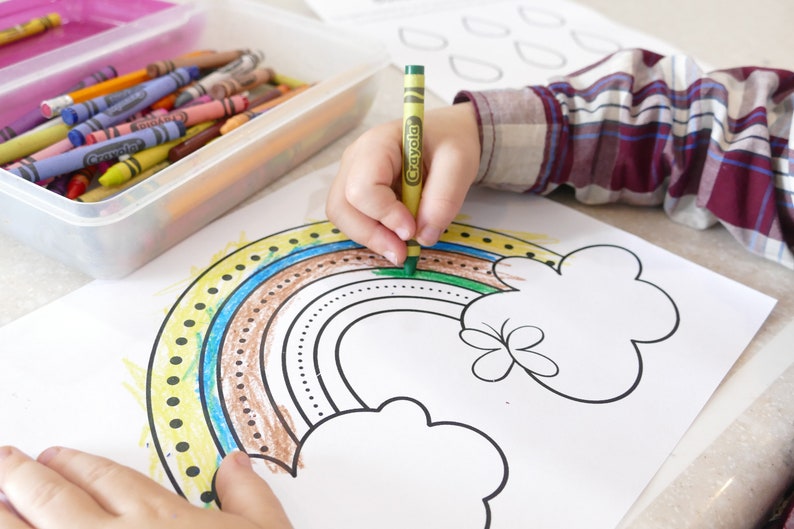 May include: A child is colouring a rainbow with crayons on a white sheet of paper. The rainbow has yellow, blue, brown, and green stripes. The child is using a green crayon to colour the top of the rainbow.
