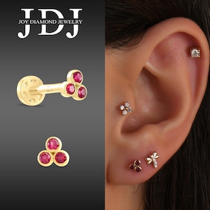 Może przedstawiać: Trzy złote kolczyki z czerwonymi kamieniami szlachetnymi. Kolczyki są pokazane na uchu osoby. Kolczyki to mały klaster trzech czerwonych kamieni szlachetnych, jeden czerwony kamień szlachetny i wzór ważki z jednym czerwonym kamieniem szlachetnym.