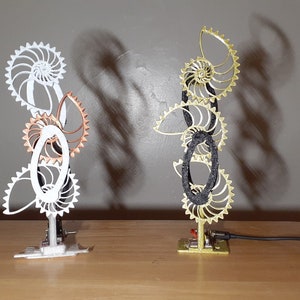 May include: Two 3D printed sculptures of a nautilus shell with intricate details. The sculpture on the left is white with copper accents, while the sculpture on the right is gold with black accents.