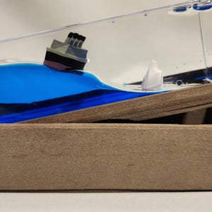 Puede incluir: Un pisapapeles rectangular de acrílico transparente con movimiento de líquido, que contiene un modelo en miniatura de un barco y un iceberg blanco en líquido azul. La pieza está inclinada sobre una base rectangular marrón texturizada.