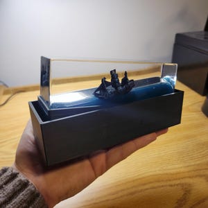Pode incluir: Um navio preto em miniatura com três mastros e figuras está envolto em uma caixa de acrílico transparente. O navio navega em uma base azul em forma de onda dentro de uma moldura retangular preta. A obra de arte é segurada em uma mão.