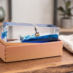 May include: A rectangular resin sculpture depicting a surfer on a wave, with a shark, inside a wooden box. The sculpture has blue and white waves, a dark blue boat, and a surfer. The box is light brown and sits on a wooden cutting board.