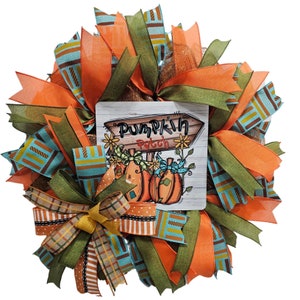 Puede incluir: Una corona de otoño con cintas naranja, verde y azul, un lazo de arpillera y un letrero que dice "Pumpkin Patch".