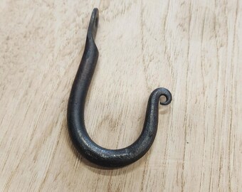 May include: A hand-forged, black metal hook with a curved design. The hook features a looped end for hanging and a decorative scroll at the bottom. The hook is shown on a light-colored wooden surface.