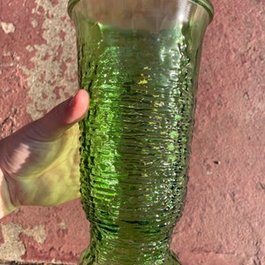 May include: A green glass vase with a textured, ridged design. The vase has a flared top and a rounded base. The glass is transparent, allowing light to pass through. The vase is approximately 9 inches tall.