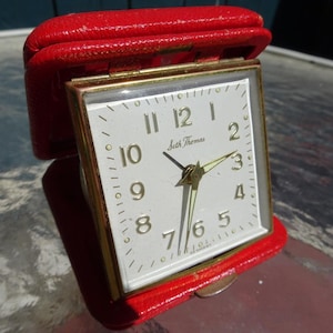 May include: A vintage Seth Thomas alarm clock with a white face and gold hands. The clock is in a red leather case.