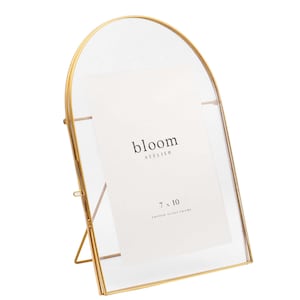 May include: A gold-coloured arched picture frame with a clear glass front. The frame holds a white paper with the text "bloom ATELIER" and "7 x 10 ARCHED GLASS FRAME". The frame has a stand on the back.