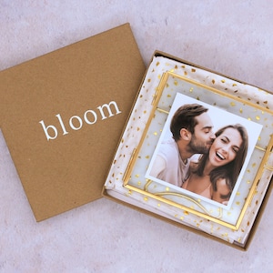 May include: A gold-toned picture frame with a white background and gold confetti. The frame contains a photo of a couple kissing. The frame is in a brown box with the word "bloom" printed on the front.