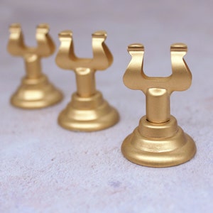 May include: Three gold metal table number holders. Each holder has a U-shaped top and a round base.