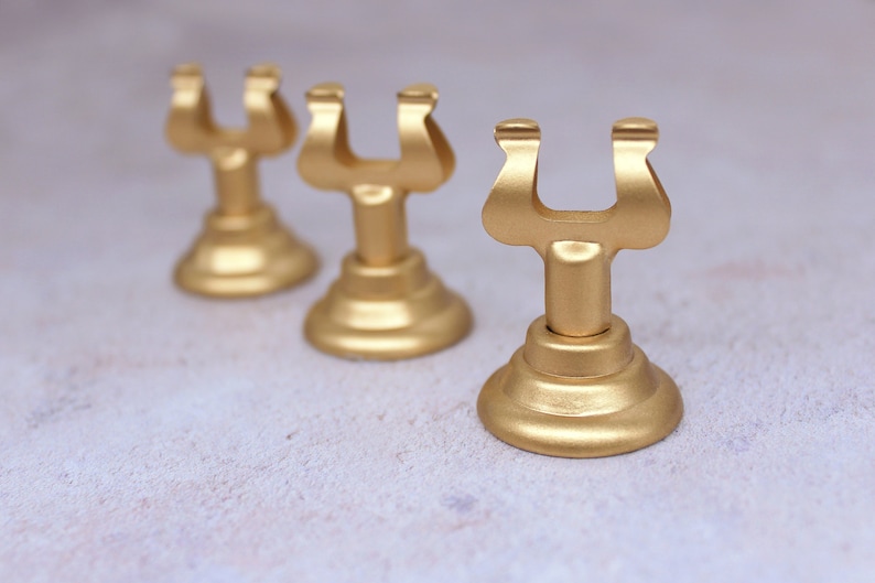 May include: Three gold metal table number holders. Each holder has a U-shaped top and a round base.
