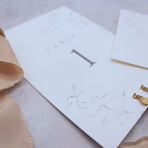 May include: A white card with the text "Table 1" and a floral design. The card is held in place by a gold metal stand.