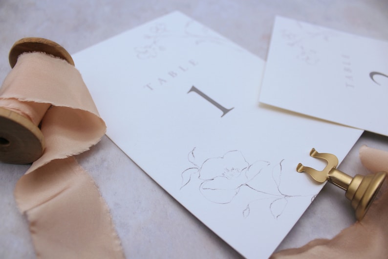 May include: A white card with the text "Table 1" and a floral design. The card is held in place by a gold metal stand.