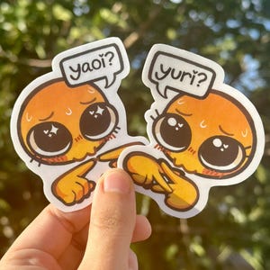 May include: Two stickers featuring orange cartoon characters with large eyes and speech bubbles. One says "yaoi?" and the other says "yuri?". The characters have yellow hands and are pointing at each other. The stickers are held in a hand.