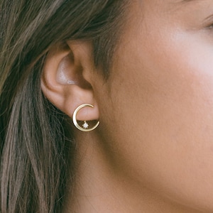 Puede incluir: Pendientes dorados en forma de media luna con una pequeña gema brillante en el centro. Los pendientes se llevan en la oreja, mostrando un diseño minimalista y celestial. Un elegante accesorio de joyería.