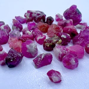 Könnte beinhalten: Eine Sammlung roher, ungeschliffener Rubin-Edelsteine in verschiedenen Rosa- und Rottönen. Die Edelsteine haben eine raue, natürliche Textur und sind auf einer weißen Oberfläche verstreut. Einige Steine weisen weiße oder graue Einschlüsse auf.