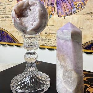 Puede incluir: Una geoda esférica con un interior cristalino de color púrpura claro que descansa sobre un soporte de pedestal de vidrio transparente facetado, junto a una torre de cristal lavanda de seis lados con una superficie rugosa y punta.