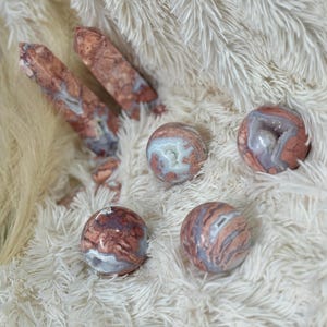 Puede incluir: Una colección de esferas de ágata pulidas y dos puntas de cristal. Las esferas tienen una mezcla de patrones rojos, blancos y grises. Las puntas de cristal son de color marrón rojizo. Los artículos se exhiben sobre una superficie blanca y esponjosa.