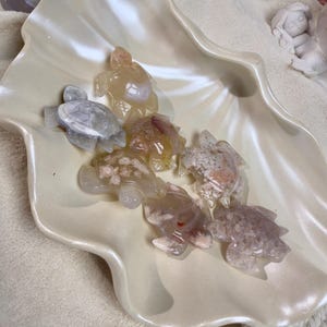 Puede incluir: Una colección de pequeñas figuras de tortugas talladas en piedras preciosas de varios colores, incluyendo blanco, gris y melocotón, se disponen en un gran cuenco nacarado con forma de concha. Las tortugas tienen detalles y se muestran sobre una superficie blanca y suave.