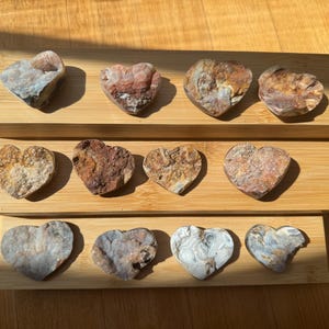 Puede incluir: Una colección de piedras en forma de corazón en varios colores, incluyendo marrón, blanco y rosa, exhibidas en tres estantes de madera. Las piedras tienen una textura rugosa y natural y están dispuestas en filas, mostrando sus patrones y tonos únicos.