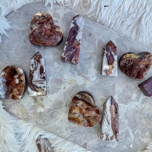 Puede incluir: Una colección de ocho cristales de ágata roja y blanca, incluyendo formas de corazón y puntiagudas. Los cristales están dispuestos sobre una superficie gris claro.