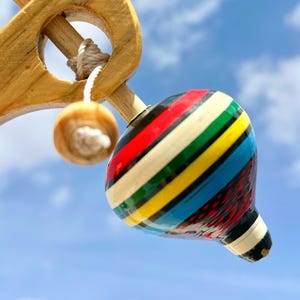 May include: A traditional wooden spinning top with a vibrant striped pattern in red, green, yellow, and blue. The toy is held by a wooden handle with a string and a small wooden ball. The background is a clear blue sky.