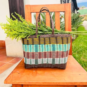 May include: A woven basket with a brown handle and a colorful striped design in yellow, teal, burgundy, and white. The basket is filled with green foliage and sits on a wooden chair.