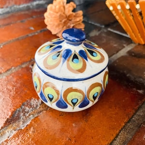 Puede incluir: Un pequeño tarro de cerámica redondo con tapa. El tarro es blanco con un patrón de diseños de plumas de pavo real azules, marrones y amarillas. La tapa es azul oscuro y tiene un pequeño pomo. El tarro está sobre una superficie de ladrillo.