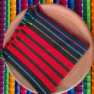 May include: A red and green woven placemat with fringe edges, sitting on a terracotta plate. The placemat has a striped pattern with a variety of colors, including red, green, yellow, blue, and white.