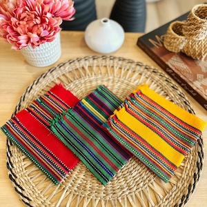 May include: Three colorful woven placemats in red, green, and yellow, each with a striped pattern. They are arranged on a round woven placemat. Decorative items, including a vase of pink flowers, are in the background.