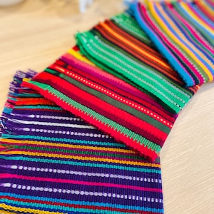 May include: Four colorful woven placemats with fringed edges. The placemats are stacked on top of each other, with the purple one on the bottom and the blue one on top. The placemats are made of a variety of colors, including purple, blue, green, red, yellow, and white.