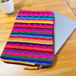 Pode incluir: Uma capa para laptop listrada colorida com um puxador de zíper de couro marrom e um zíper marrom. A capa é feita de tecido tecido e tem um padrão de arco-íris.