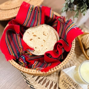 May include: A woven basket with a lid, filled with a flatbread and a red and multi-colored woven fabric. The basket is sitting on a woven tray with a wooden sign that reads "Home Sweet Home".