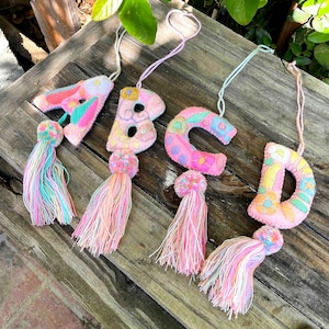 May include: Four hanging ornaments shaped like the letters A, B, C, and D. Each letter is crafted from pink felt with colorful floral embroidery and attached to a matching yarn tassel. Each ornament features a string loop for hanging purposes.
