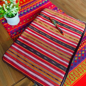 Pode incluir: Uma capa para laptop com um padrão listrado colorido em tons de vermelho, laranja, rosa e marrom. A capa tem um fecho de correr e um puxador de couro castanho.