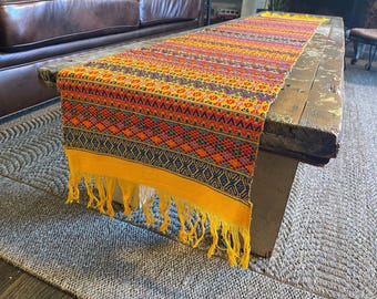Op de afbeelding: Een gele tafelrunner met een kleurrijk geometrisch patroon en franjes. De runner ligt op een houten tafel met een verweerde afwerking.
