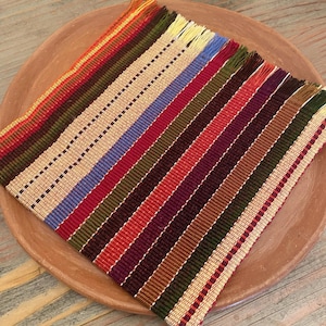 May include: A colorful, striped woven placemat with fringed edges rests on a terracotta plate. The placemat features a variety of colors, including red, blue, green, and beige, arranged in vertical stripes. The plate is round and has a slightly raised edge.