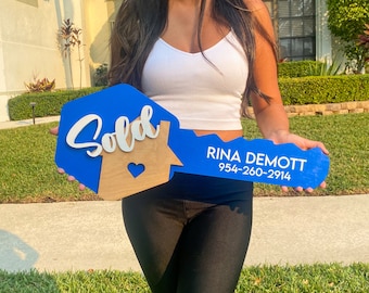 May include: A large, blue wooden key-shaped sign with the word "Sold" in white script. A wooden house shape with a heart cutout is on the key. The name "Rina Demott" and a phone number are printed on the key.