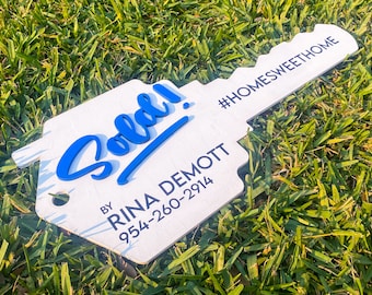 May include: A white wooden key-shaped sign with blue text that reads "Sold!" and "By Rina Demott 954-260-2914" and "#HOMESWEETHOME".
