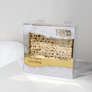 May include: A clear acrylic box with a gold-colored accent, holding matzah. The box has a gold-colored inscription in Hebrew and English, with the words "Carin Family" visible. The matzah is a light brown color.