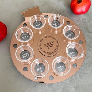 May include: A copper-colored seder plate with eight small glass bowls and a central inscription. The plate is shaped like a pomegranate and includes Hebrew text. Two red pomegranates are visible in the background.